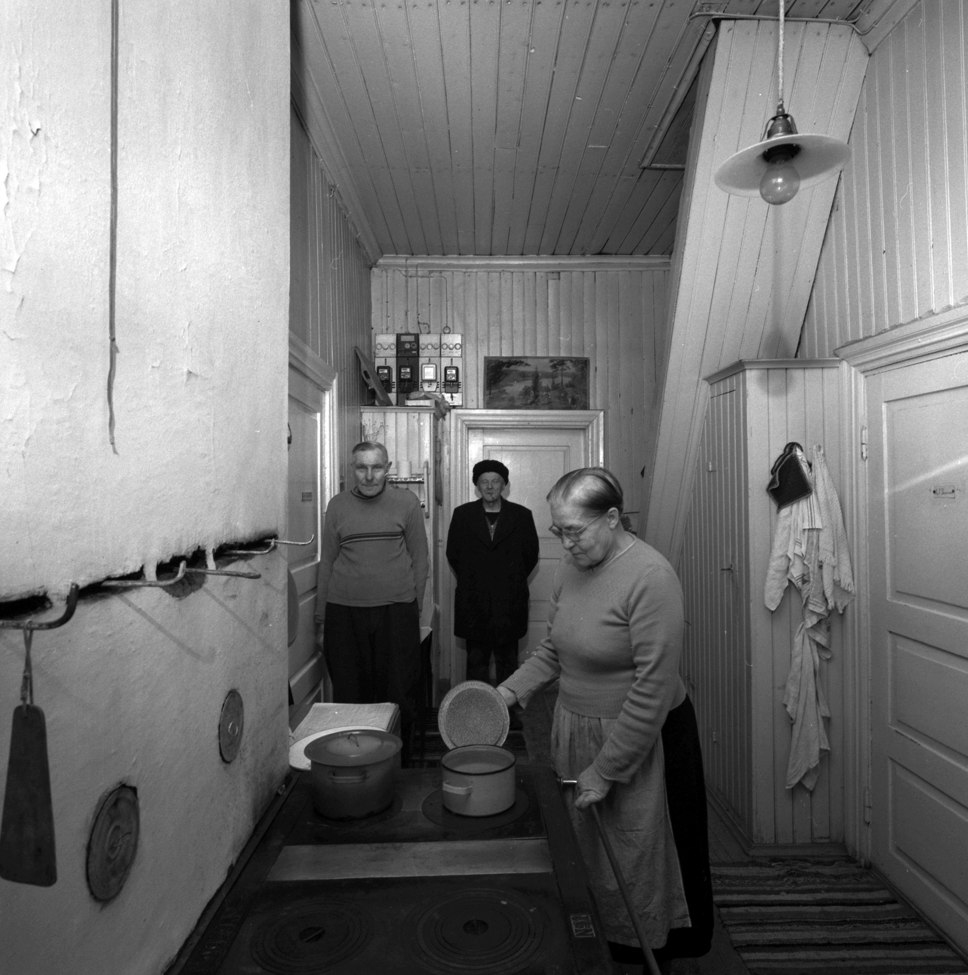 Kolme ihmistä seisoo yhteiskäyttöisessä keittiötilassa. Puuhellan vieressä seisovalla naisella on kädessään hellalla olevan kattilan kansi.