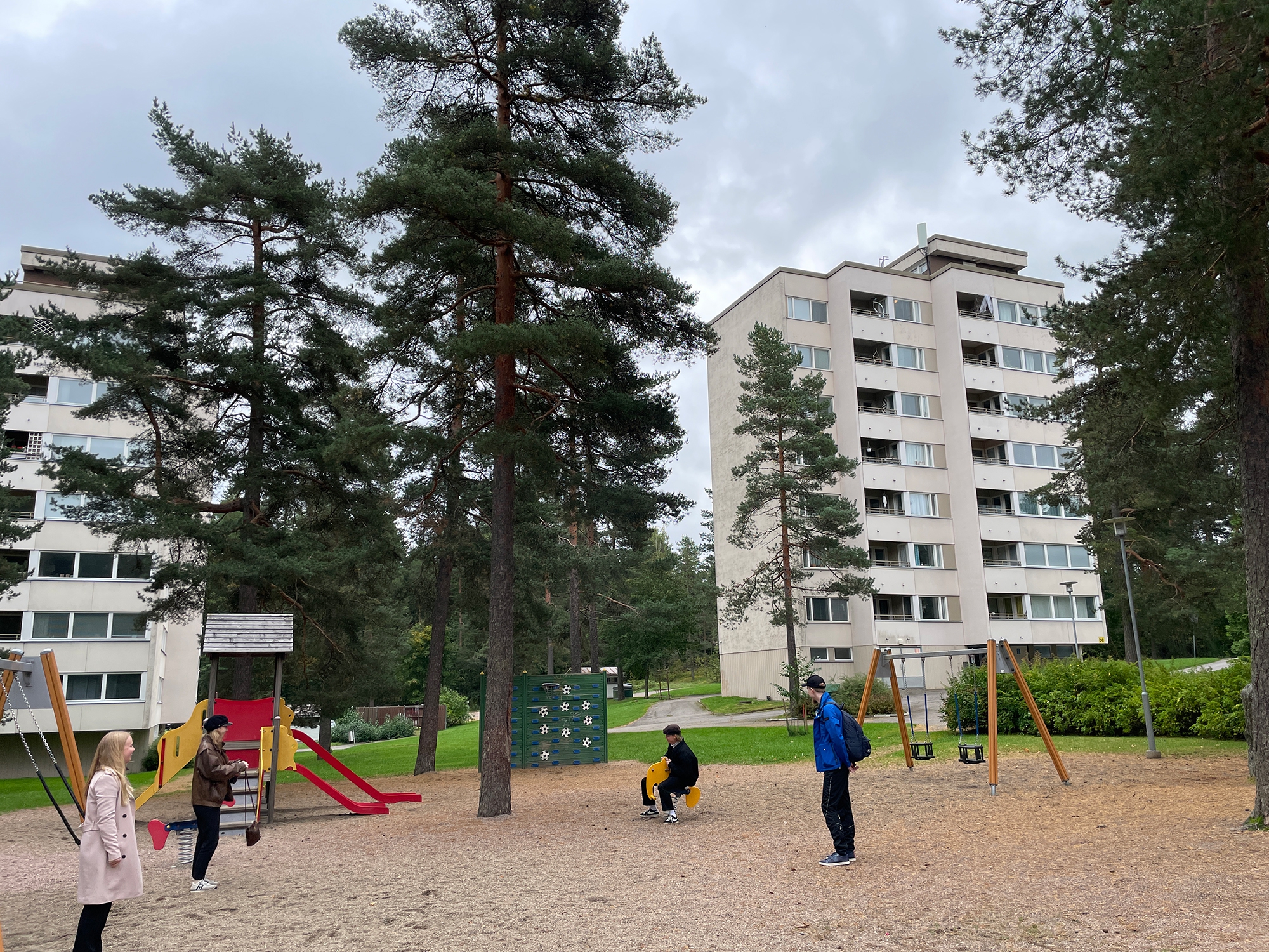 Neljä aikuista seisoo etualan leikkipuistossa keinujen ja liukumäen vieressä. Taustalla puistomaisessa ympäristössä kaksi vaaleaa kahdeksankerroksista, yhden rapun asuinkerrostaloa.