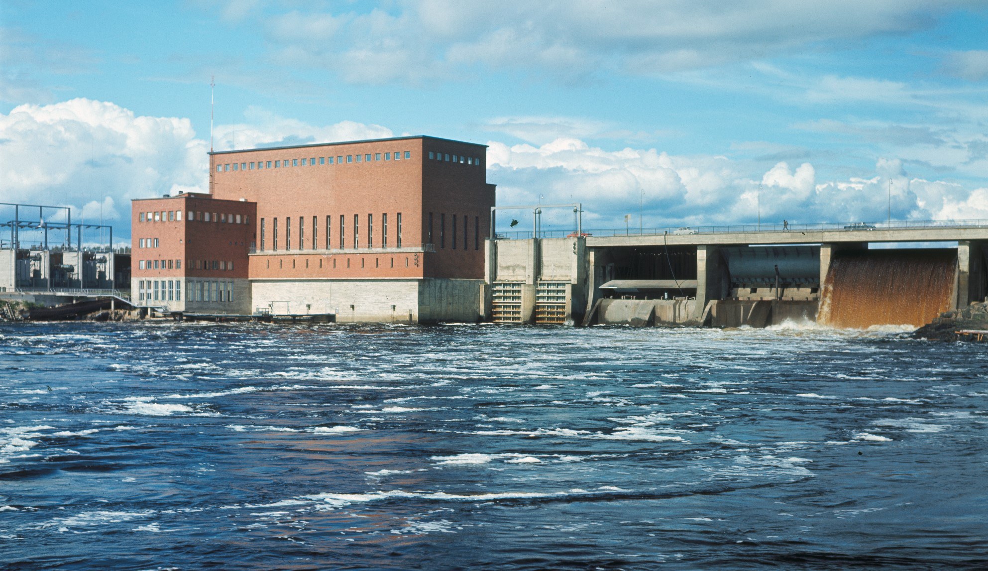 Yleiskuva voimalaitoksesta. Vasemmalla muuntajakenttää, keskellä punatiilinen voimalaitosrakennus, oikealla betonisia patorakenteita sulkuportteineen. Kuvan etualalla alaveden virtauksia.