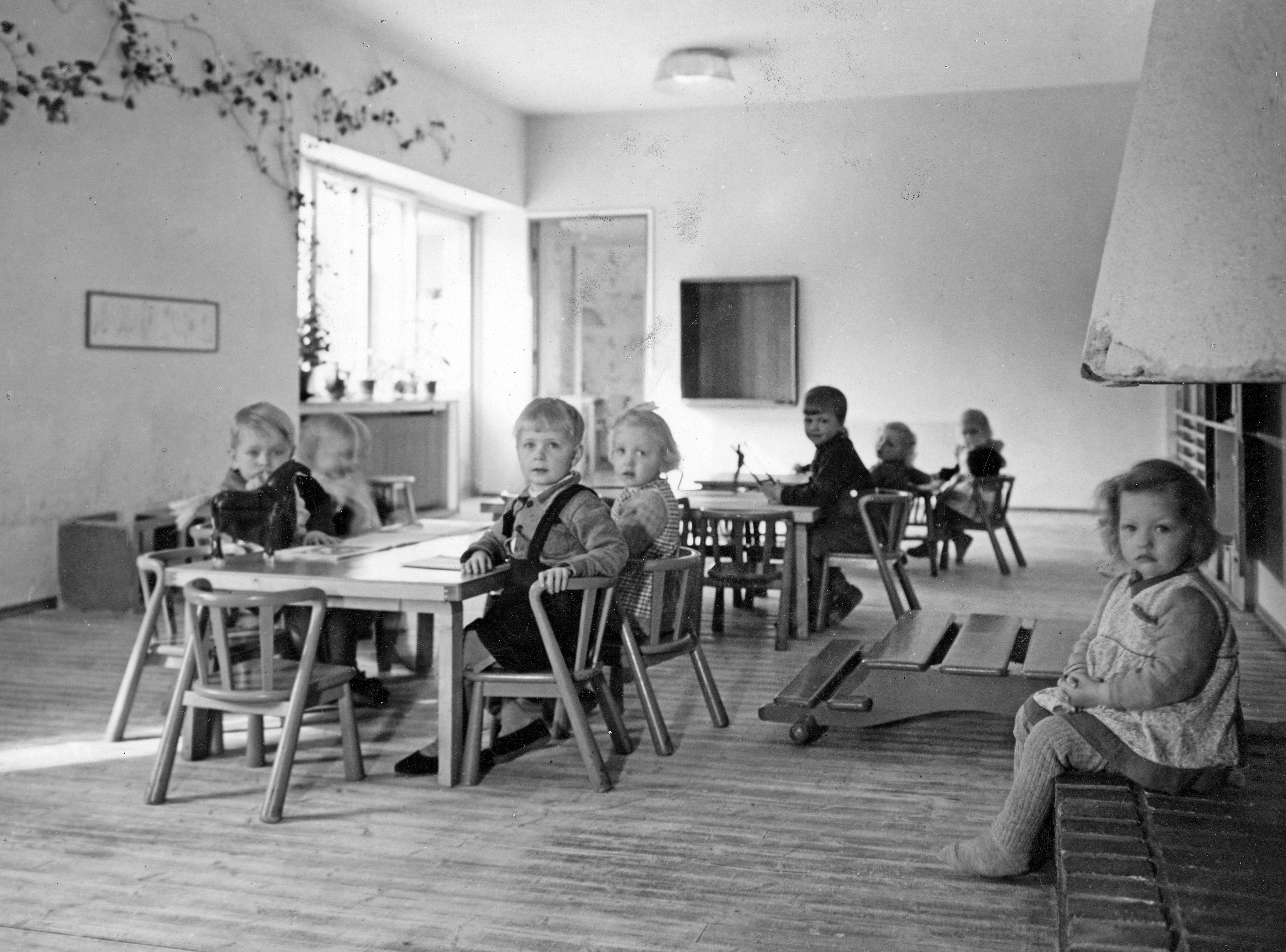 Mustavalkoisessa valokuvassa kahdeksan päiväkoti-ikäistä lasta istuu kameraan katsoen pienten pöytien ääressä valoisassa huonetilassa. Yksi lapsista istuu vasemmalla olevan takan reunalla.
