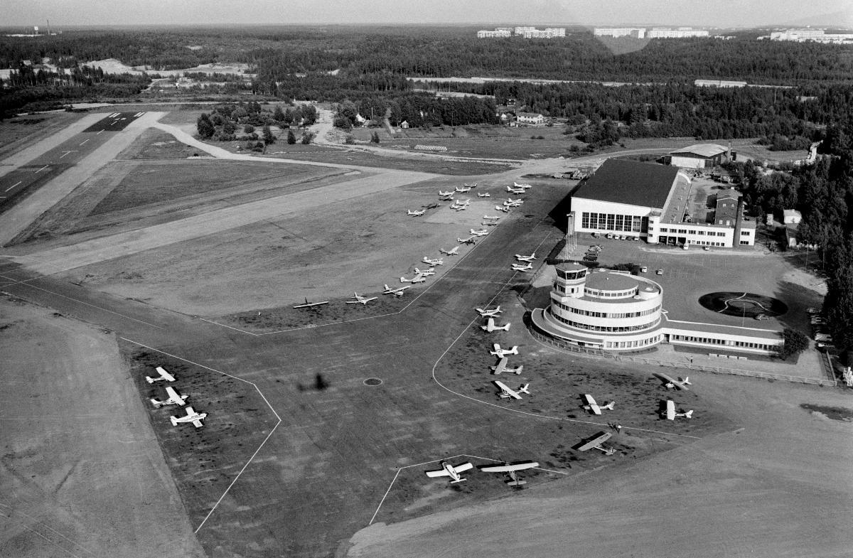 Viistoilmakuva lentokenttäalueesta, jonka vasemmalla laidalla ovat kiitotiet ja oikealla laidalla terminaali- sekä hangaarirakennukset. Kentällä pysäköitynä pienkoneita. Taustalla on metsää ja kaukana horisontissa kerrostaloja.