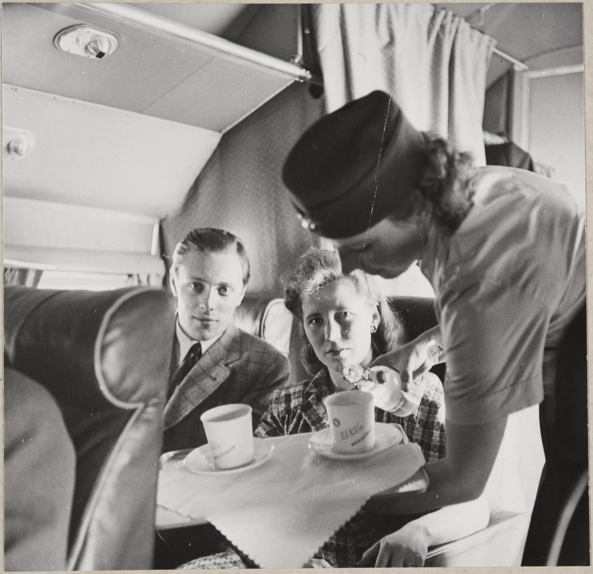 Kuva lentokoneen matkustamosta. Kaksi matkustajaa istumassa. Lentoemäntä kaataa juomaa mukiin.