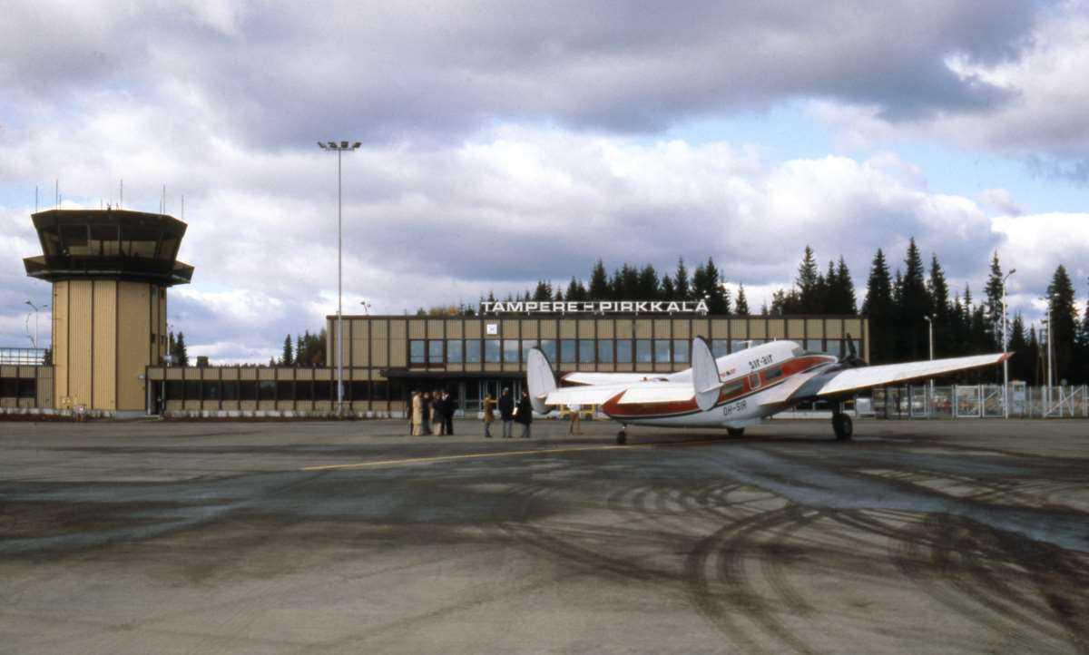 Näkymä kenttäalueen suunnasta lentokenttäterminaalille, jonka katolla teksti on Tampere-Pirkkala. Vasemmassa reunassa on lennonjohtotorni. Kentällä olevan lentokoneen vieressä seisoo muutamia ihmisiä.
