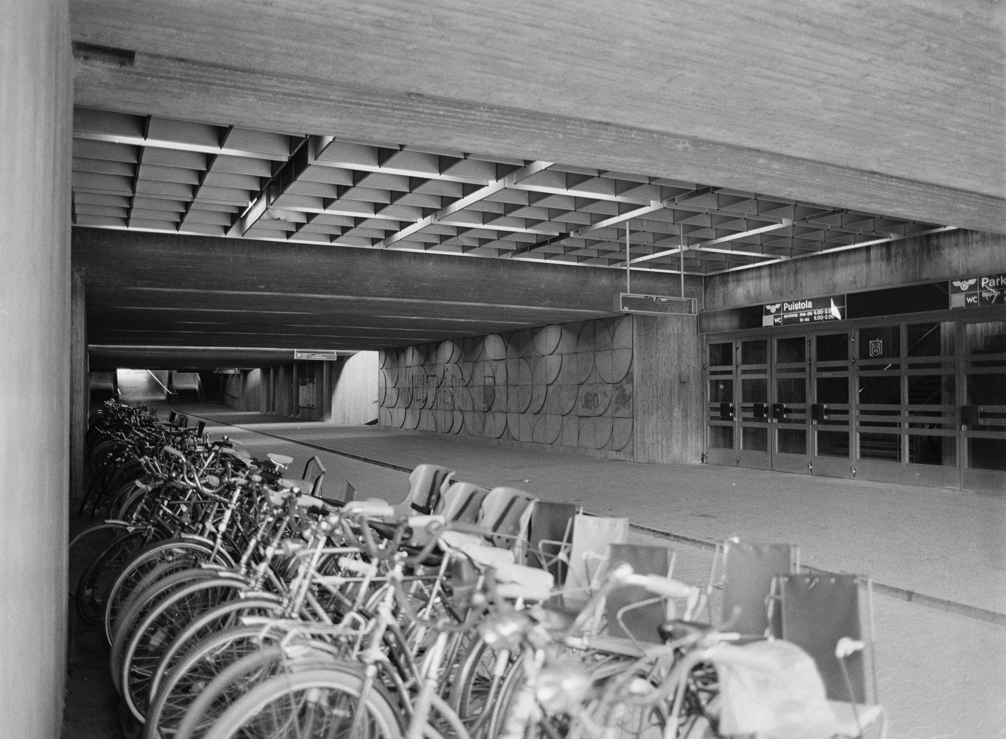 Kuva radan alikulkukäytävästä, jonka vasemmassa reunassa polkupyöriä. Oikealla puolella betoniseinässä reliefi ja rivi ovia. Katossa betonipalkit ja ruudutettu vyöhyke ovien kohdalla.