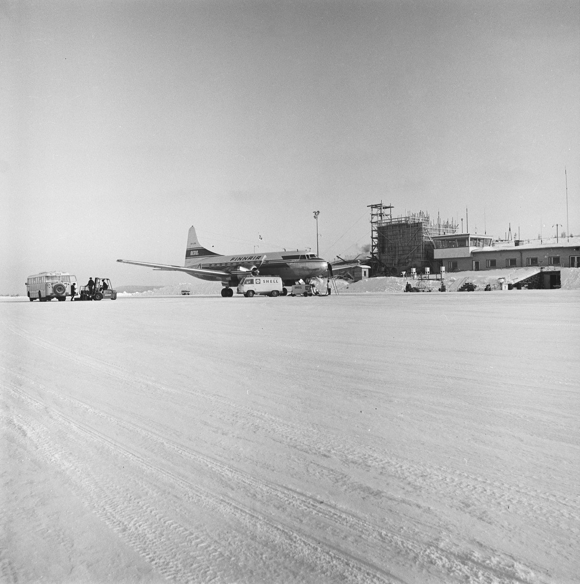 Talvinen näkymä, jossa lentokone kentällä tankattavana. Koneen edessä on pakettiauto ja läheisyydessä linja-auto sekä matkalaukkuja siirtävä ajoneuvo. Taustalla matala lentoasemarakennus, josta osa on vielä rakenteilla.