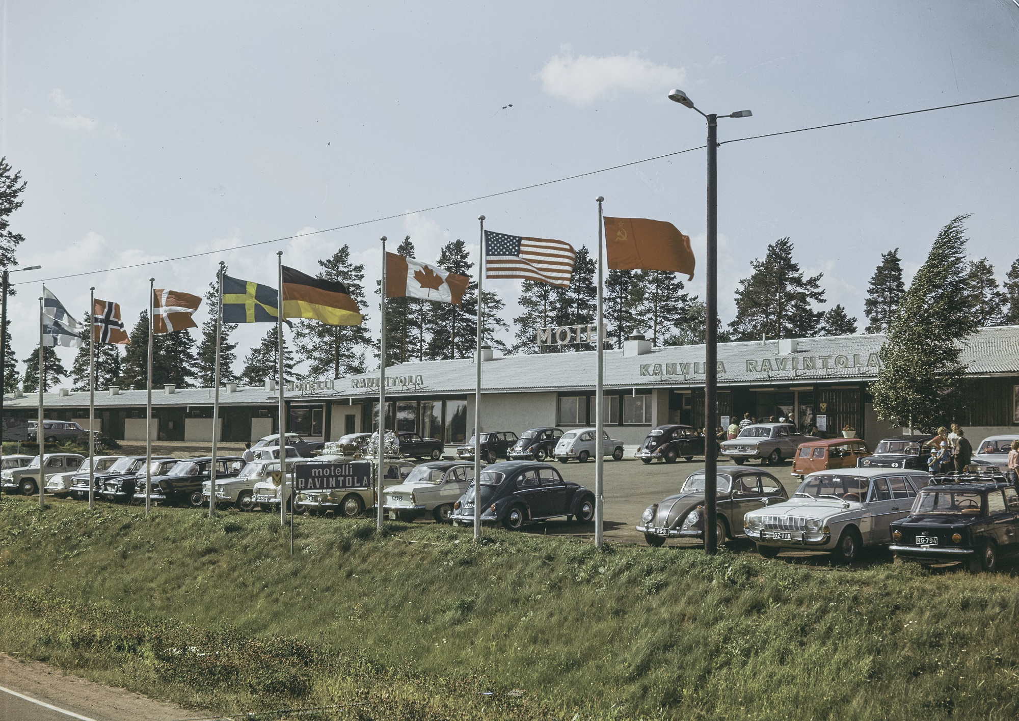 Näkymä motellialueelle. Etualalla nurmipenkereen päällä on kahdeksan lipputankoa, jossa liehuu eri maiden lippuja. Niiden takana paikoitusalue, jossa on pysäköitynä lukuisia henkilöautoja. Yksikerroksinen motellirakennus reunustaa parkkialuetta.