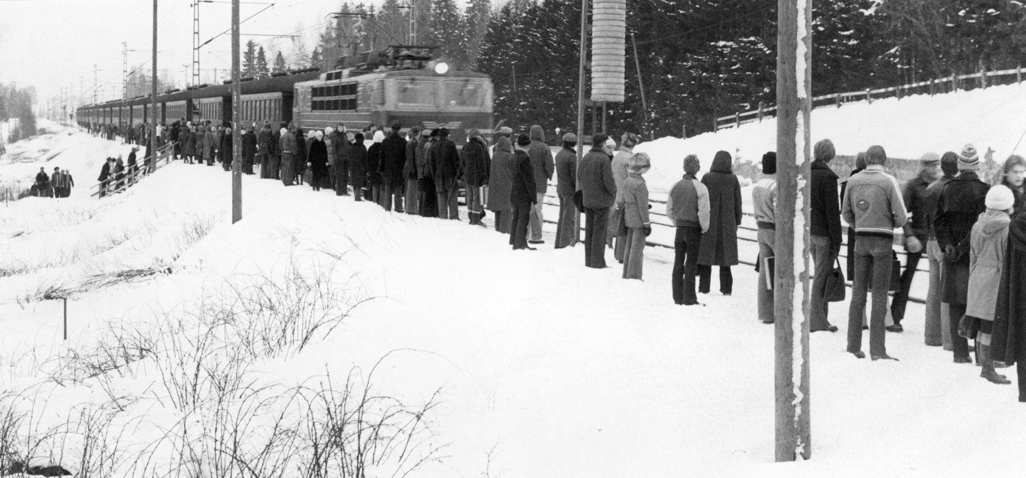 Talvinen näkymä. Juna saapumassa seisakkeelle, jossa seisoo runsaasti ihmisiä.