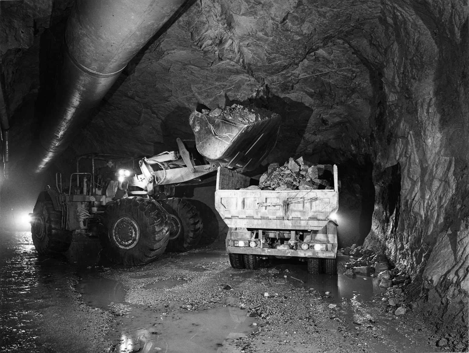 Kauhakuormaaja lastaa kuorma-autoa maa-aineksella kallioon louhitun tunnelin levennyksessä. Taustalla siintää valo ja tunnelin katossa kulkee putki.