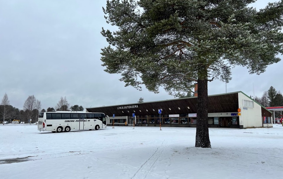Värikuvassa asemarakennus lumisena päivänä. Rakennuksen edessä on bussi. Etualalla tuuhea mänty.