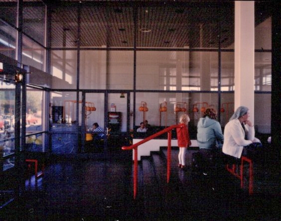 Värikuvassa aseman sisätila, jonka keskellä lasiseinä. Etualalla ihmisiä istumassa penkeillä.